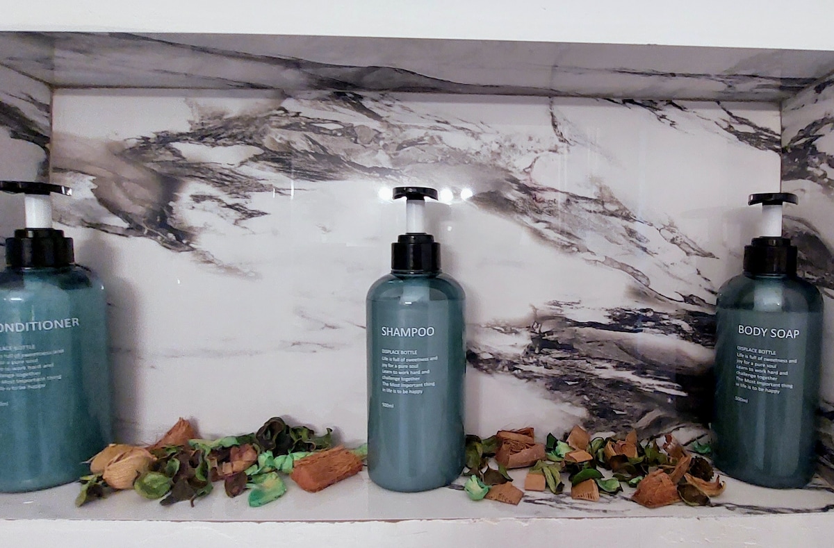 Three refillable bottles labeled Conditioner, Shampoo, and Body Soap are neatly displayed on a marble shelf. The bottles feature a dark teal color, and a mixture of natural materials, including dried leaves and wood, is arranged at the base, enhancing the modern aesthetic.