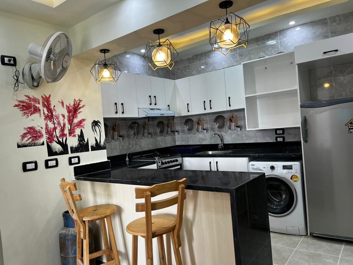 A modern kitchen space features a black countertop with two wooden bar stools. Wall-mounted lights highlight the sleek cabinetry and appliances, including a washing machine and gas stove. Decorative elements, such as wall art in vibrant colors, add an aesthetic touch.