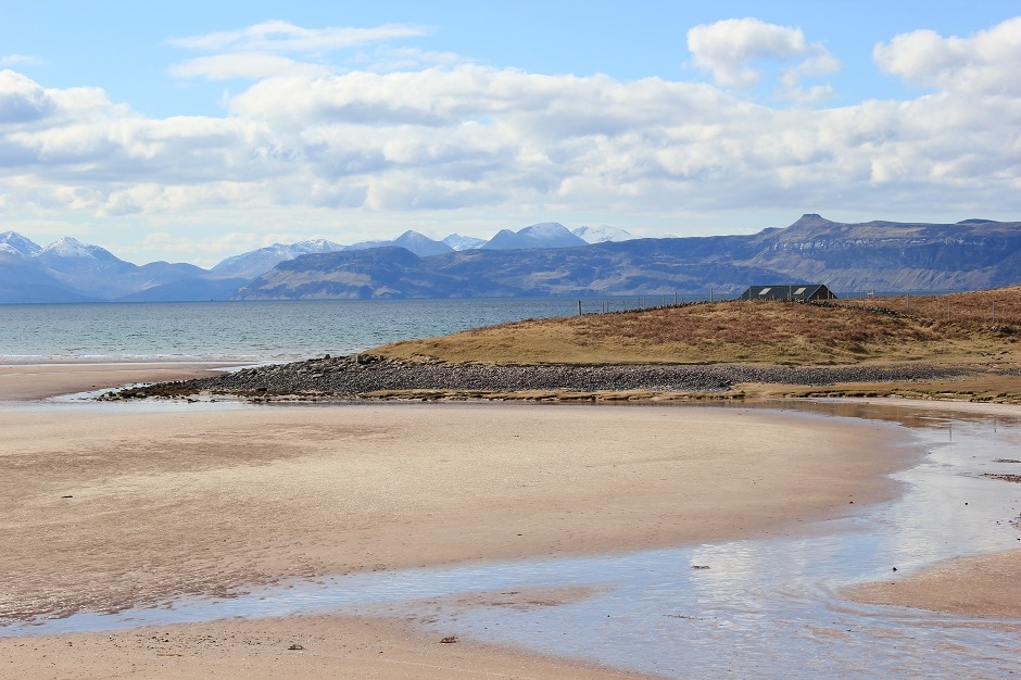 A serene landscape showcases a sandy shore with gentle curves, leading to a calm body of water. The distant mountains provide a scenic backdrop, partially covered with snow. Soft clouds drift across a clear sky, creating a peaceful ambiance in the area.