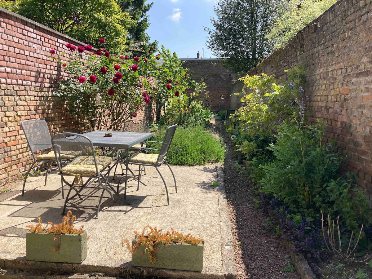 A well-maintained garden area features a stone patio with a metal dining set, surrounded by colorful flowering plants and lush greenery. A low wall made of brick encloses the space, providing a sense of privacy. The sunny atmosphere enhances the inviting setting.
