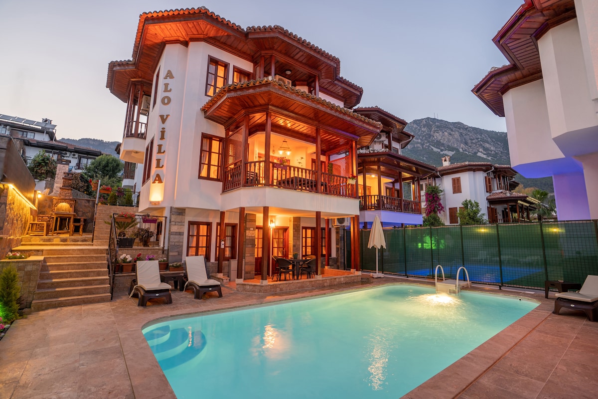A multi-level villa is depicted with a charming wooden balcony on each floor. The inviting private pool reflects the evening light, surrounded by sun loungers. Lush greenery complements the outdoor space, with mountains visible in the backdrop.