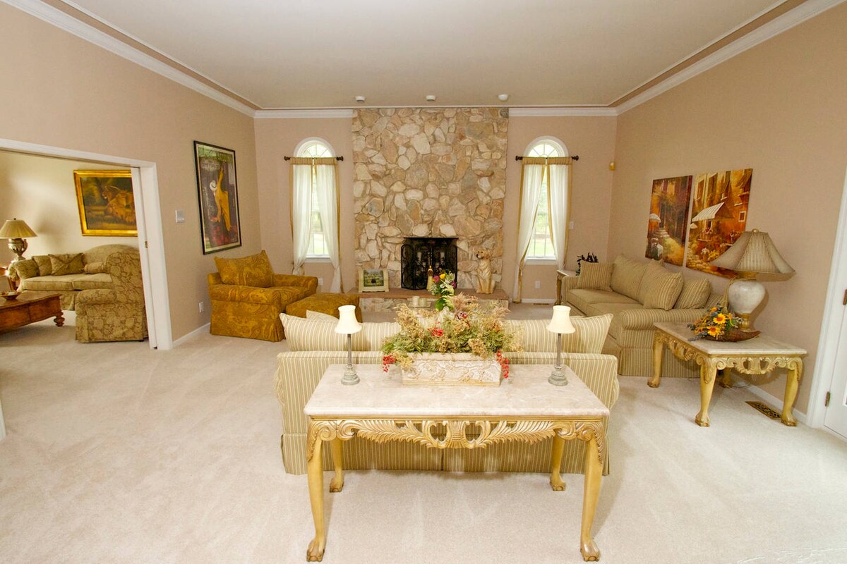 A spacious living area features a large stone fireplace as a focal point, flanked by elegant armchairs and couches. Natural light enters through arched windows adorned with light drapes. Decorative tables with floral arrangements add a touch of warmth to the inviting setting.