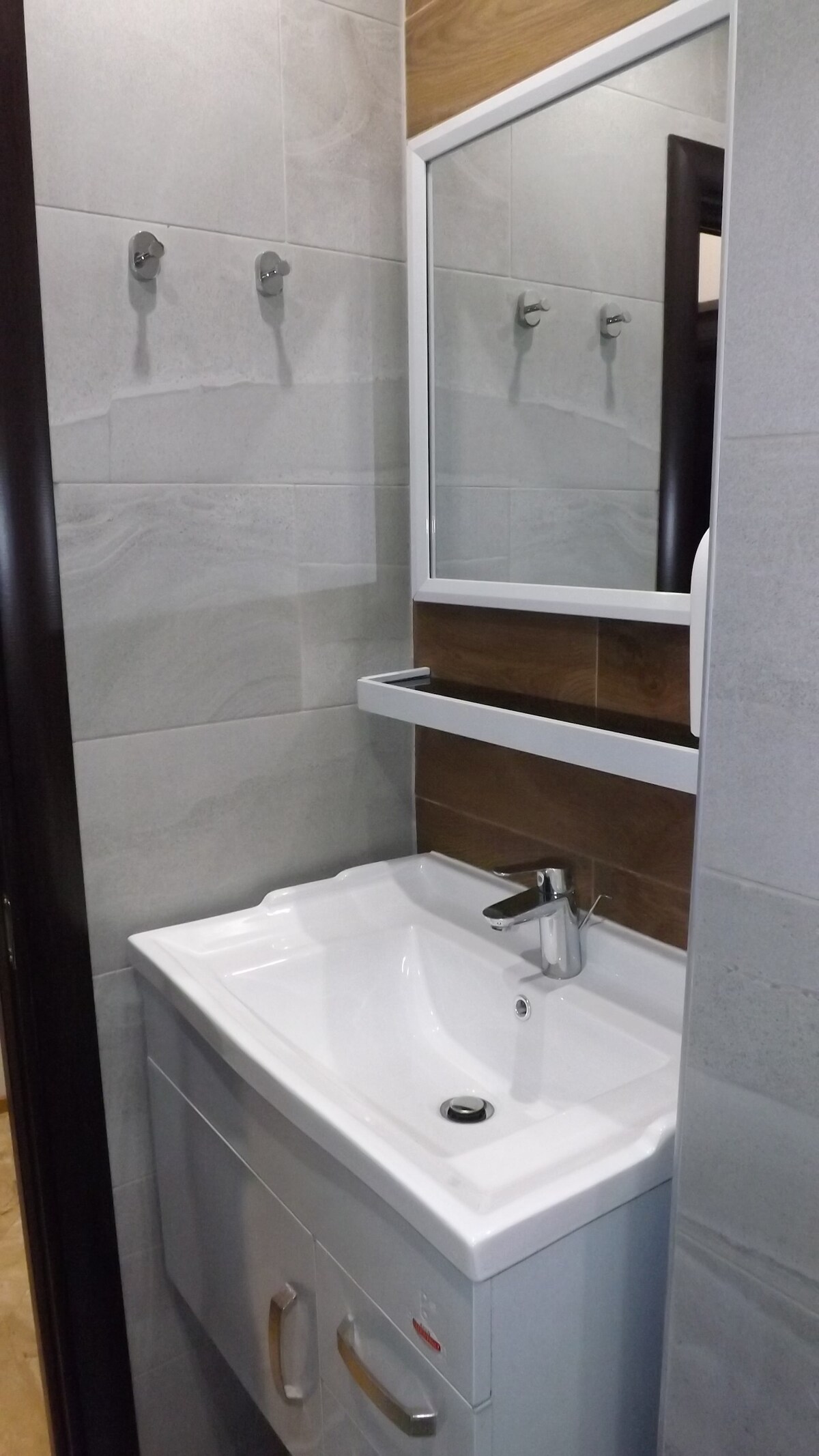 A modern bathroom features sleek gray tiles and a spacious vanity with a white sink. A mirror is mounted above the sink, reflecting the minimalist design. Two hooks are offered on the wall for hanging towels or personal items.