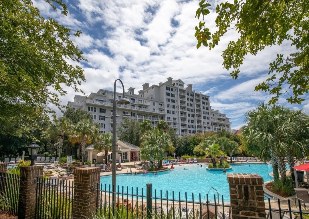 The image displays a large, inviting pool area surrounded by lush palm trees and greenery. Sun loungers are positioned beside the pool, and a multi-story condominium building is visible in the background under a partly cloudy sky.