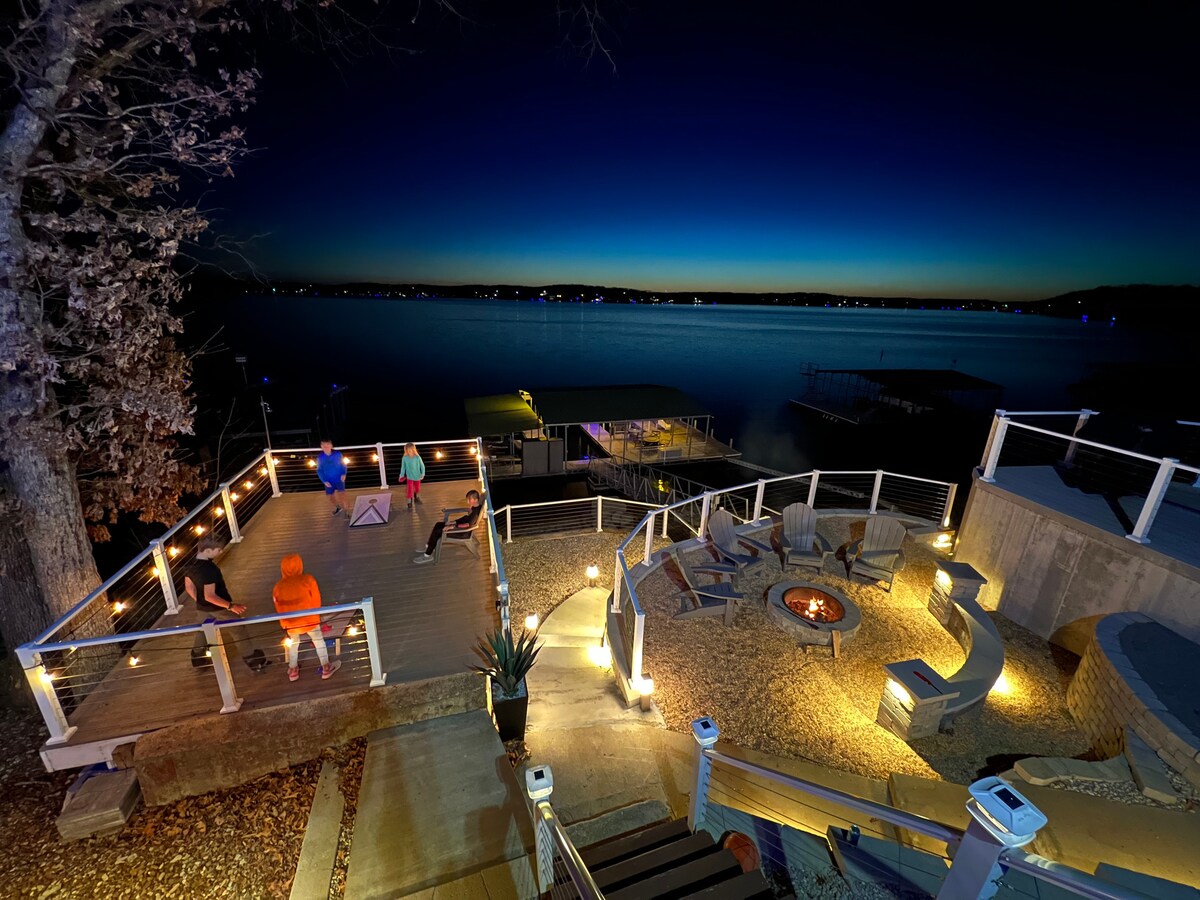 An outdoor space is highlighted at dusk, featuring a fire pit surrounded by seating and a wraparound deck. Soft ambient lighting enhances the tranquil lakeview, with the horizon displaying a gradient of deep blue to light blue. Nearby watercraft and distant lights reflect off the water.