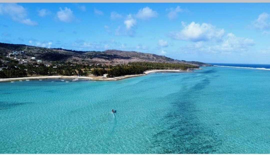 A clear turquoise lagoon reflects the sky's soft clouds, with gentle waves lapping at the shoreline. A small boat is seen in the distance, surrounded by the shimmering blue water. Lush green hills rise in the background, framing the tranquil coastal scene.