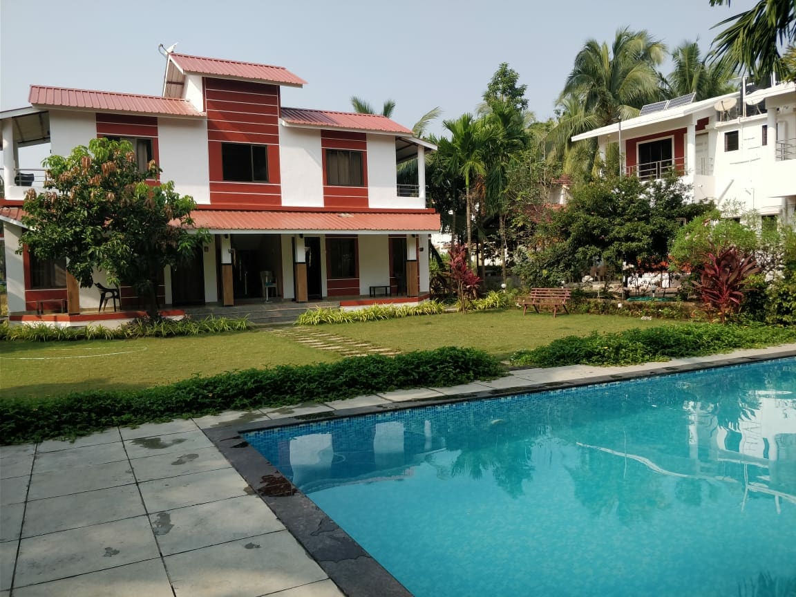 The image captures a two-story cottage surrounded by lush greenery and well-maintained gardens. A clear swimming pool is visible in the foreground, with a stone patio leading to the structured outdoor area, featuring shaded seating and vibrant foliage enhancing the tranquil setting.