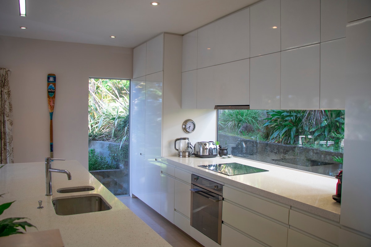 A modern kitchen is highlighted by sleek white cabinetry and a spacious countertop. Stainless steel appliances are visible, including a stovetop and kettle. Natural light filters in through an adjacent window, illuminating the greenery outside.