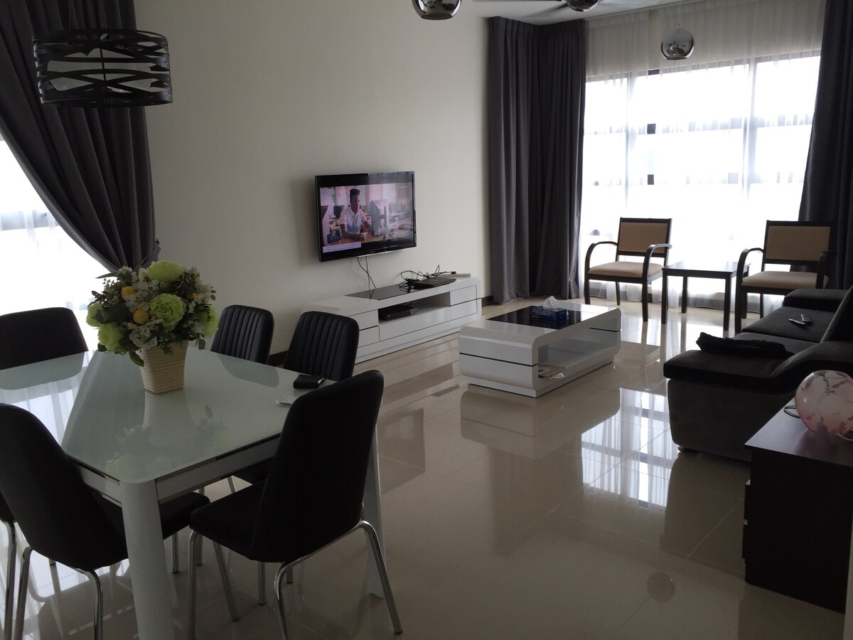 A modern living area features a sleek white dining table surrounded by black chairs. A flat-screen TV is mounted on the wall, with a stylish coffee table positioned nearby. Elegant curtains frame large windows, allowing natural light to brighten the room.