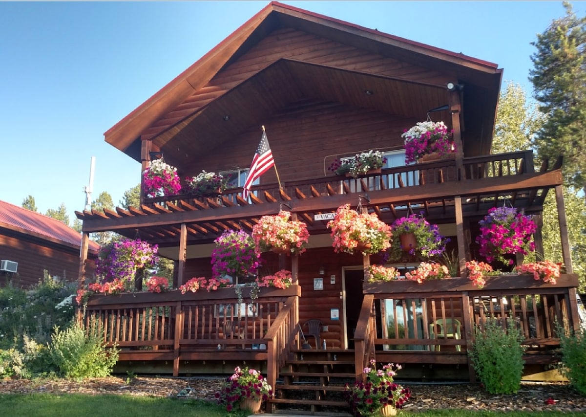 The cabin's exterior is adorned with vibrant flower arrangements, enhancing the inviting front porch. An American flag is displayed prominently, welcoming guests. The two-story wooden structure features a spacious deck surrounded by lush greenery and well-maintained landscaping.