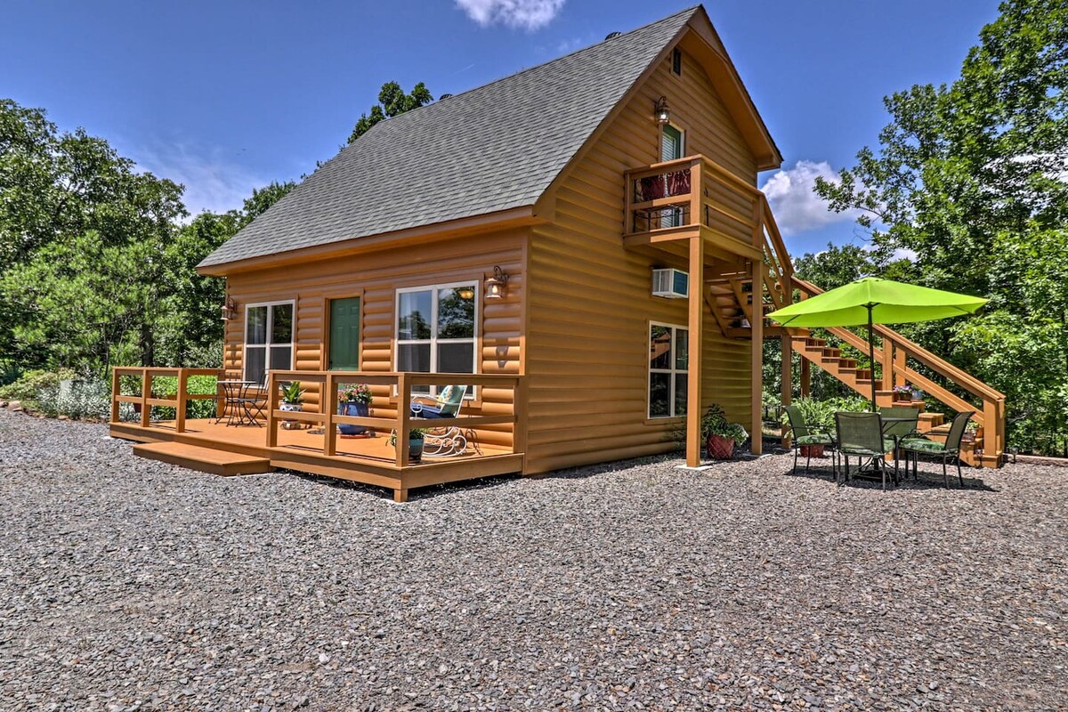 A two-story guesthouse with a rustic wooden exterior is surrounded by a gravel area. Spacious decks feature seating and a small dining set under a bright umbrella. Lush greenery is visible in the background, complementing the vibrant blue sky.