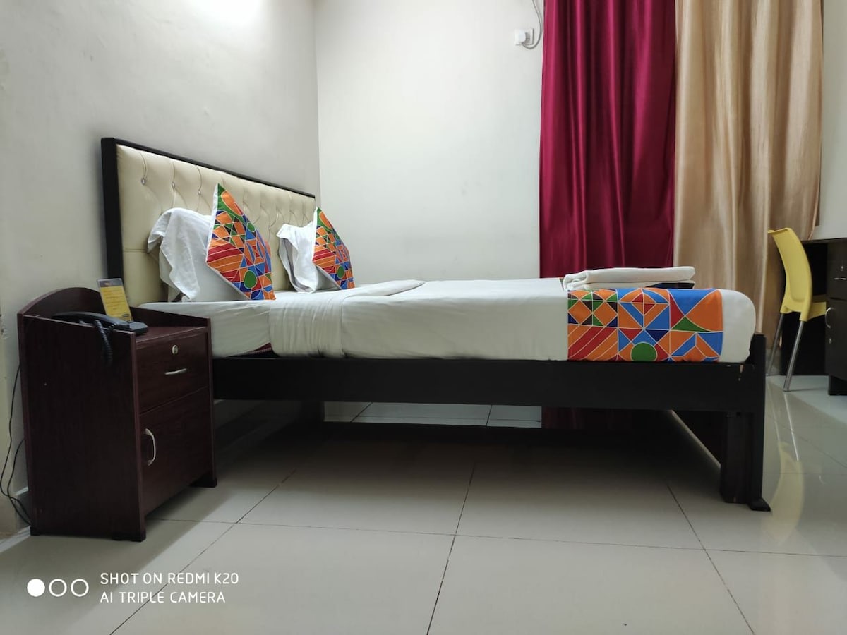 A spacious bed is positioned against a neutral wall, adorned with brightly patterned cushions. Soft lighting highlights the bed's elegant headboard, while curtains in warm hues frame the window. A bedside table and chair are visible, contributing to the overall functional layout.