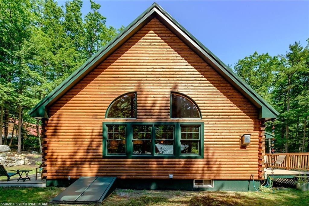 The cabin's exterior features a rustic wooden facade with large arched windows that allow natural light to fill the interior. A welcoming deck extends from the house, surrounded by lush greenery and mature trees, providing a serene woodland setting.