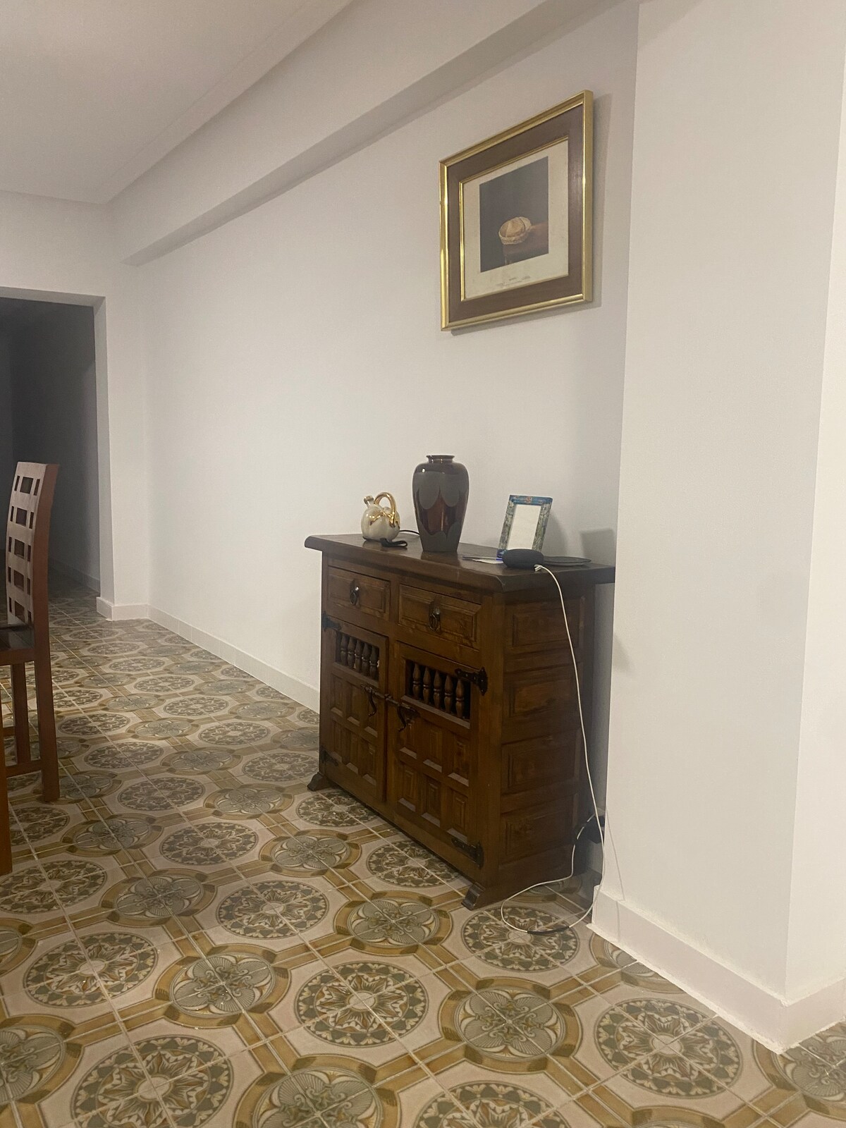 A wooden cabinet is positioned against a white wall, adorned with decorative items such as a vase and framed artwork. The floor features intricate patterned tiles, contributing to the room's character. A small electronic device is placed on the cabinet's surface.