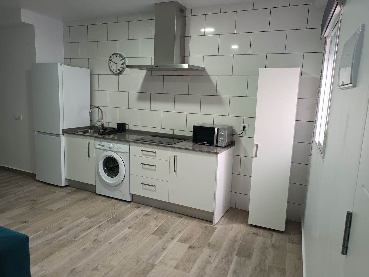 The kitchen area features modern appliances, including a refrigerator, washing machine, and microwave. It is characterized by a sleek design with white cabinetry and a neutral tiled backsplash. Natural light enters through a nearby window, illuminating the functional workspace.