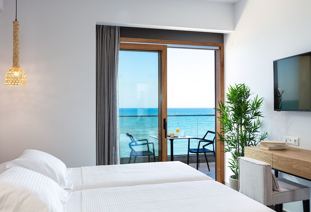 A bright studio room features two spacious beds with white linens. Floor-to-ceiling glass doors open to a private balcony overlooking the Mediterranean sea. A small table and two chairs are positioned outside, complemented by a decorative indoor plant. A wall-mounted television and minimalist desk complete the space.