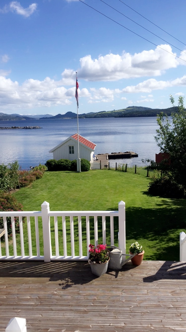 A well-maintained garden with green grass leads towards a calm fjord. A small, white wooden house with a red roof is positioned beside a dock. The distant hills create a serene backdrop under a partly cloudy sky. Flowerpots add a touch of color to the patio.