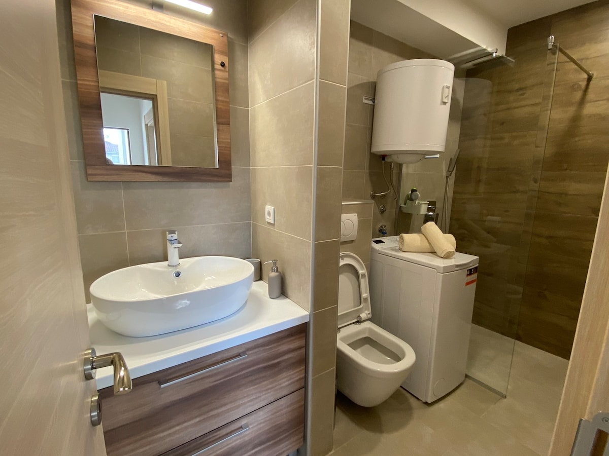A modern bathroom features a combination of neutral tiles and wooden accents. A circular sink is positioned atop a wooden cabinet, with a large mirror above. Essential amenities, including a washing machine, are visible, alongside a shower area enclosed in glass.