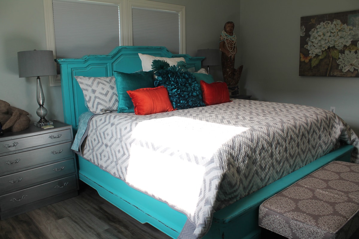 A vibrant bedroom features a spacious bed adorned with a textured grey and white blanket, bright orange and turquoise decorative pillows, and a teal headboard. Adjacent are two bedside lamps and a silver dresser, while natural light filters through the partially covered windows.