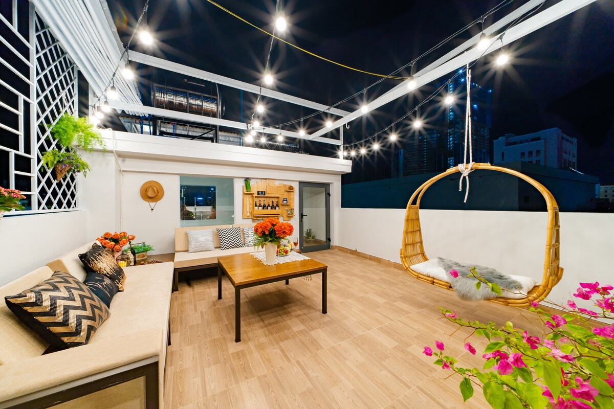 A spacious rooftop patio is illuminated with string lights, featuring a cozy seating area with a soft upholstered couch and a wooden coffee table. A hanging chair adds a unique touch, while potted plants and vibrant flowers enhance the welcoming environment.