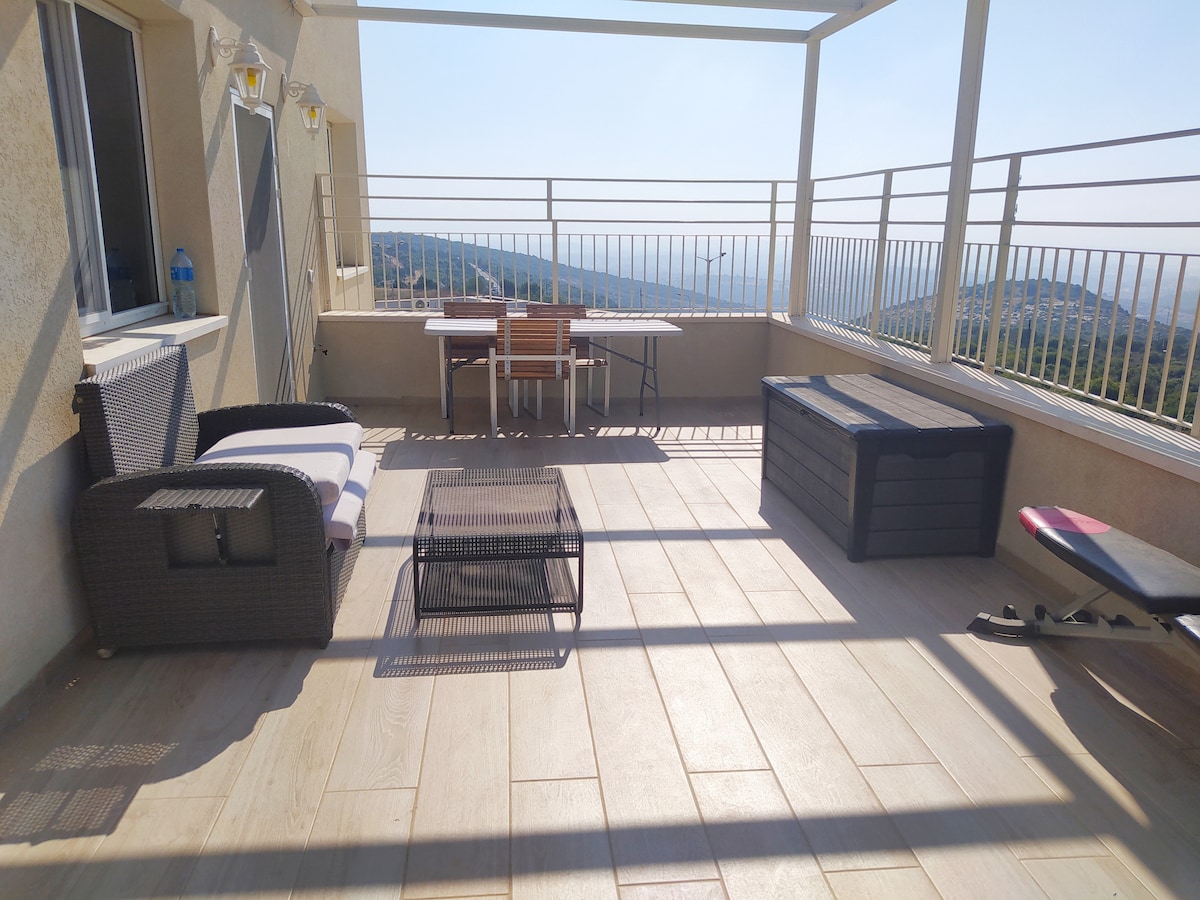 A spacious outdoor porch is featured, offering uninterrupted views of Haifa Bay and the Galilee hills. Outdoor furniture includes a seating area with a small table, lounge chairs, and a storage box, all set on light-colored flooring. The atmosphere is bright and airy.