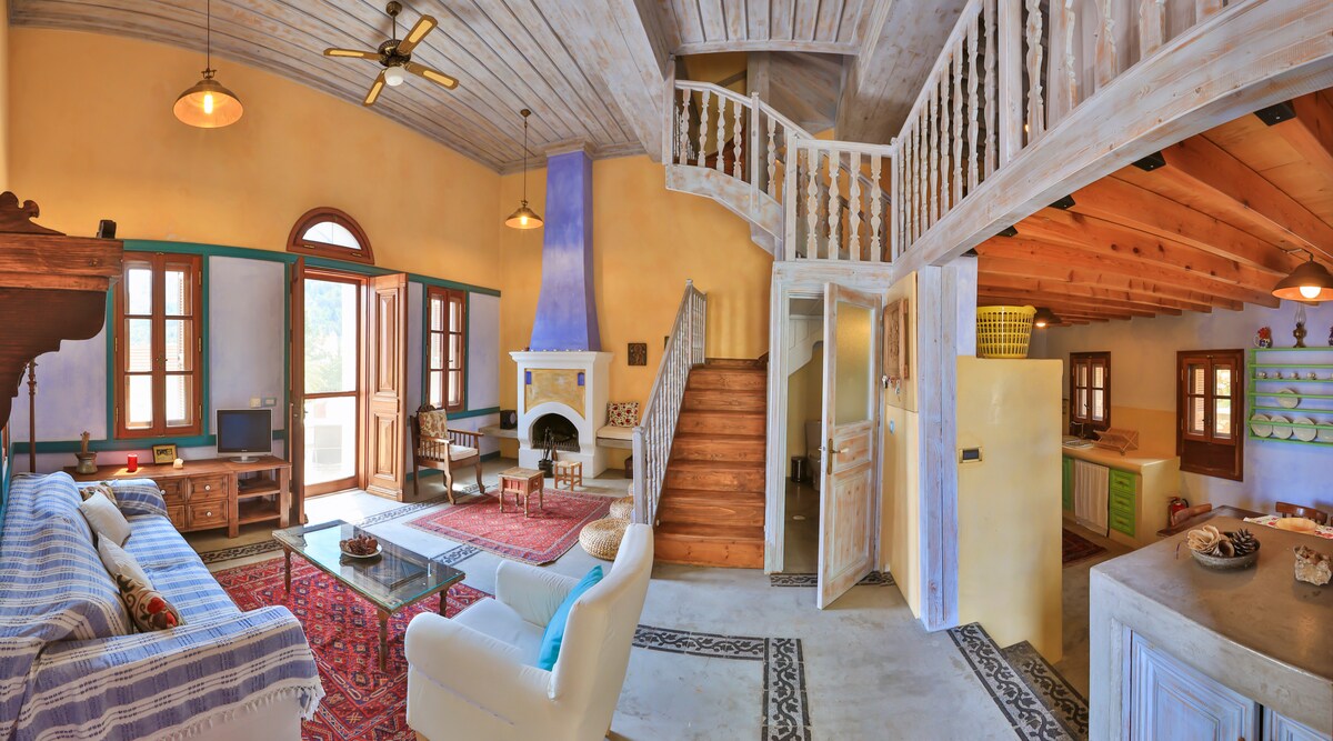 An open living space features a blend of traditional and modern design elements. Large windows allow natural light to fill the room. A comfortable white sofa and coffee table offer seating, while a staircase leads to the upper level. Bright walls complement wooden beams and decorative rugs.