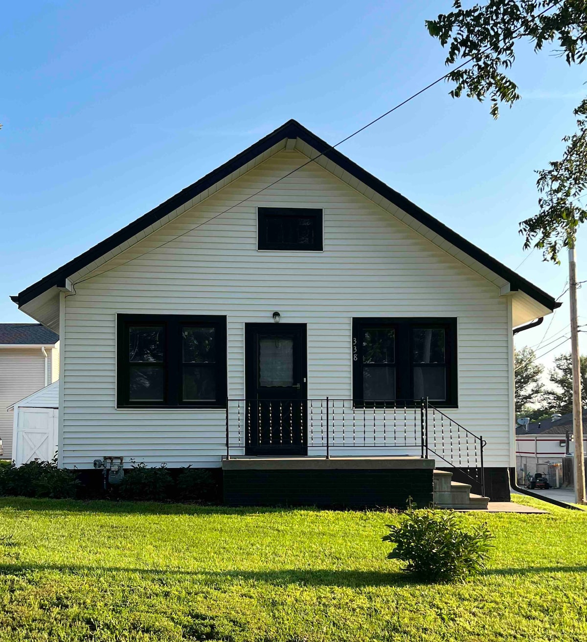 The exterior of a charming cottage showcases a white facade with black trim. A black door is framed by an inviting porch, featuring decorative railings. Lush green grass surrounds the entrance, while trees provide gentle shade in the background.