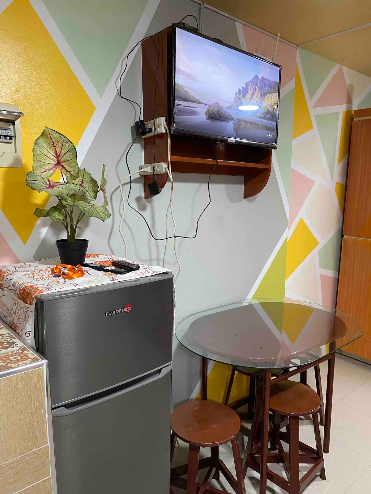 A compact kitchen area features a modern refrigerator and a glass-topped dining table with three wooden chairs. A television is mounted on the wall above the table. The walls exhibit a colorful geometric design with pastel shades, creating a lively atmosphere.