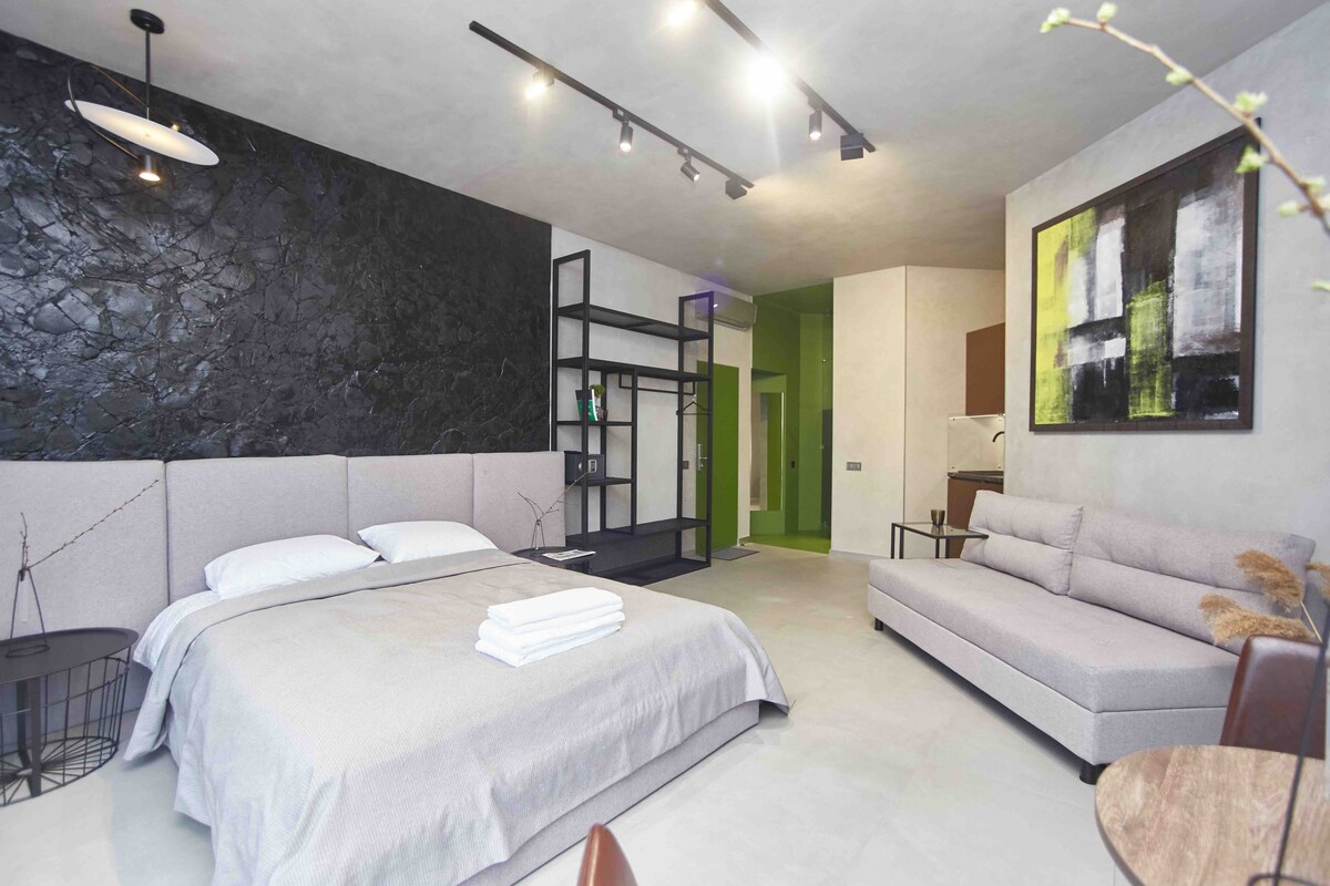 A modern studio apartment features a spacious bed with neatly arranged towels. A minimalist black accent wall contrasts with muted tones, while a light grey sofa is positioned nearby. Open shelving adds functionality, and a small kitchenette is visible in the background.