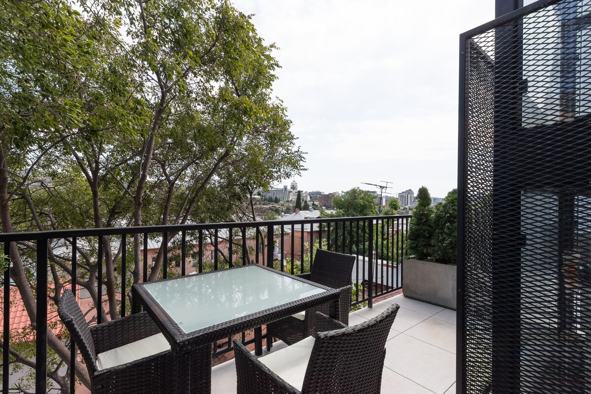 An outdoor terrace features a glass-topped table surrounded by four wicker chairs. The railing showcases a distant view of rooftops and buildings under a cloudy sky, while trees provide a natural backdrop.