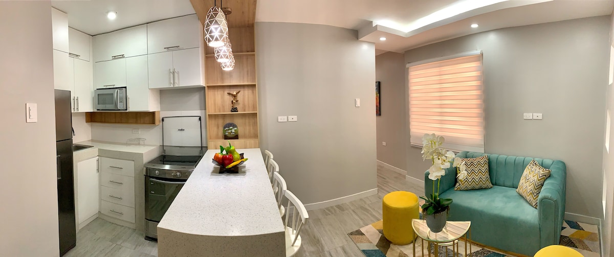 An open concept space is highlighted, featuring a modern kitchen and a cozy living area. Sleek white cabinetry and stainless steel appliances complement the gray tones. A turquoise sofa with decorative cushions, a yellow accent table, and a dining bar with stools create an inviting environment.