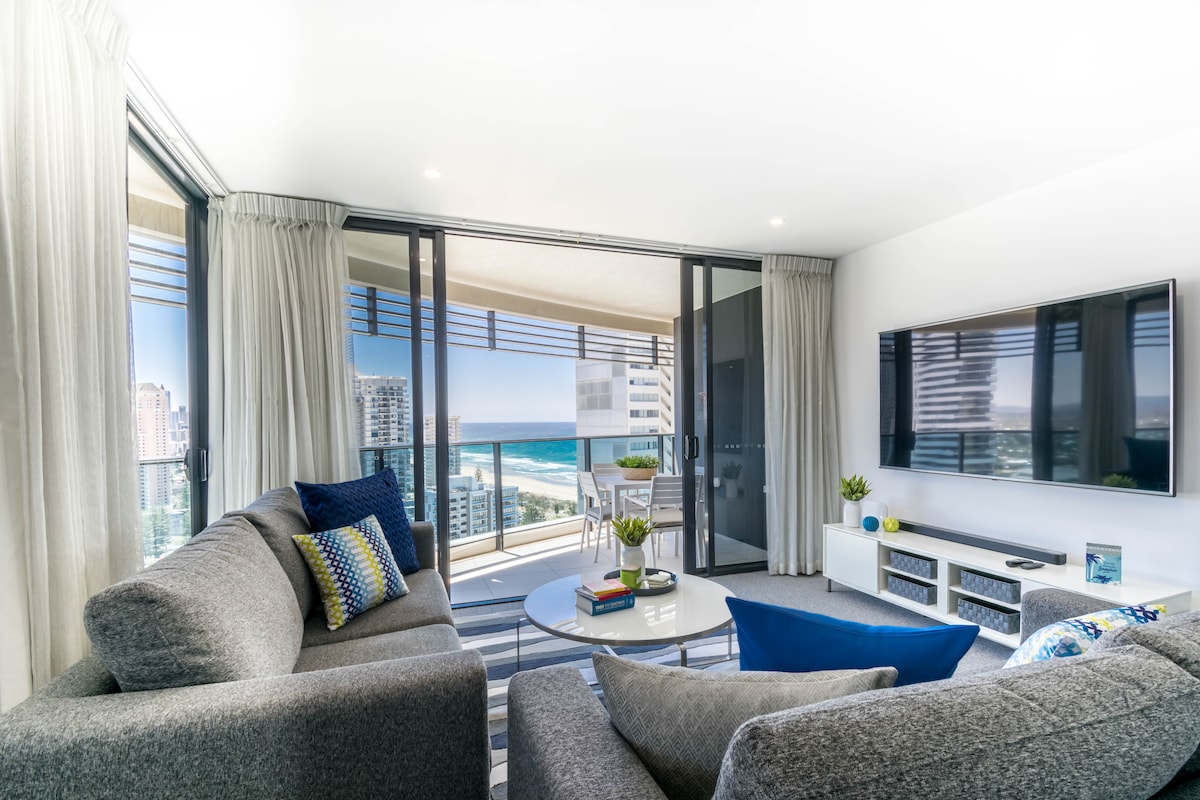 A cozy living area features a comfortable grey sofa set and a TV mounted on the wall. Large glass doors lead to a wrap-around balcony with ocean views, while natural light fills the space through sheer curtains.