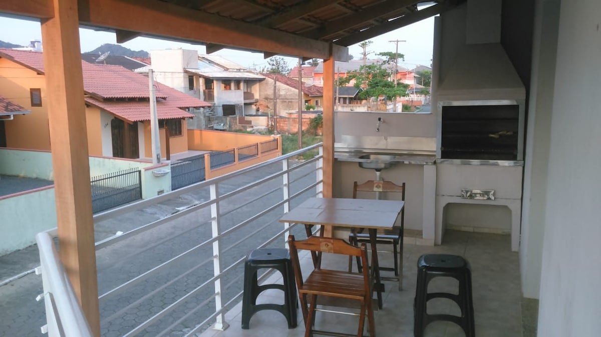 An outdoor balcony area is visible, featuring a barbecue grill and a small dining table with four chairs. The space is partially shaded and offers views of the neighborhood, including nearby houses and greenery. The floor is tiled for easy maintenance.