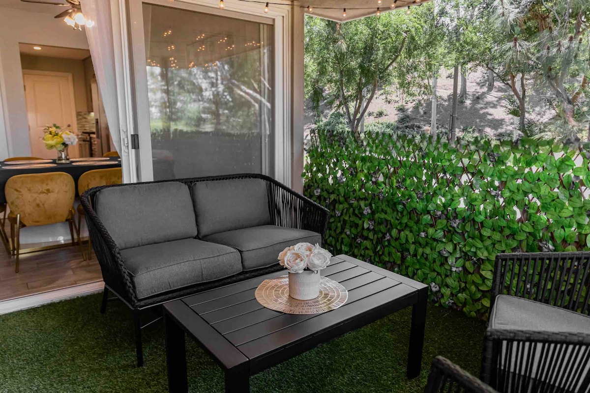 A comfortable seating area is shown, featuring a black wicker couch and matching armchairs. A dark coffee table rests in front, adorned with a vase of flowers. Surrounding greenery provides a sense of privacy, while string lights add a gentle glow overhead.