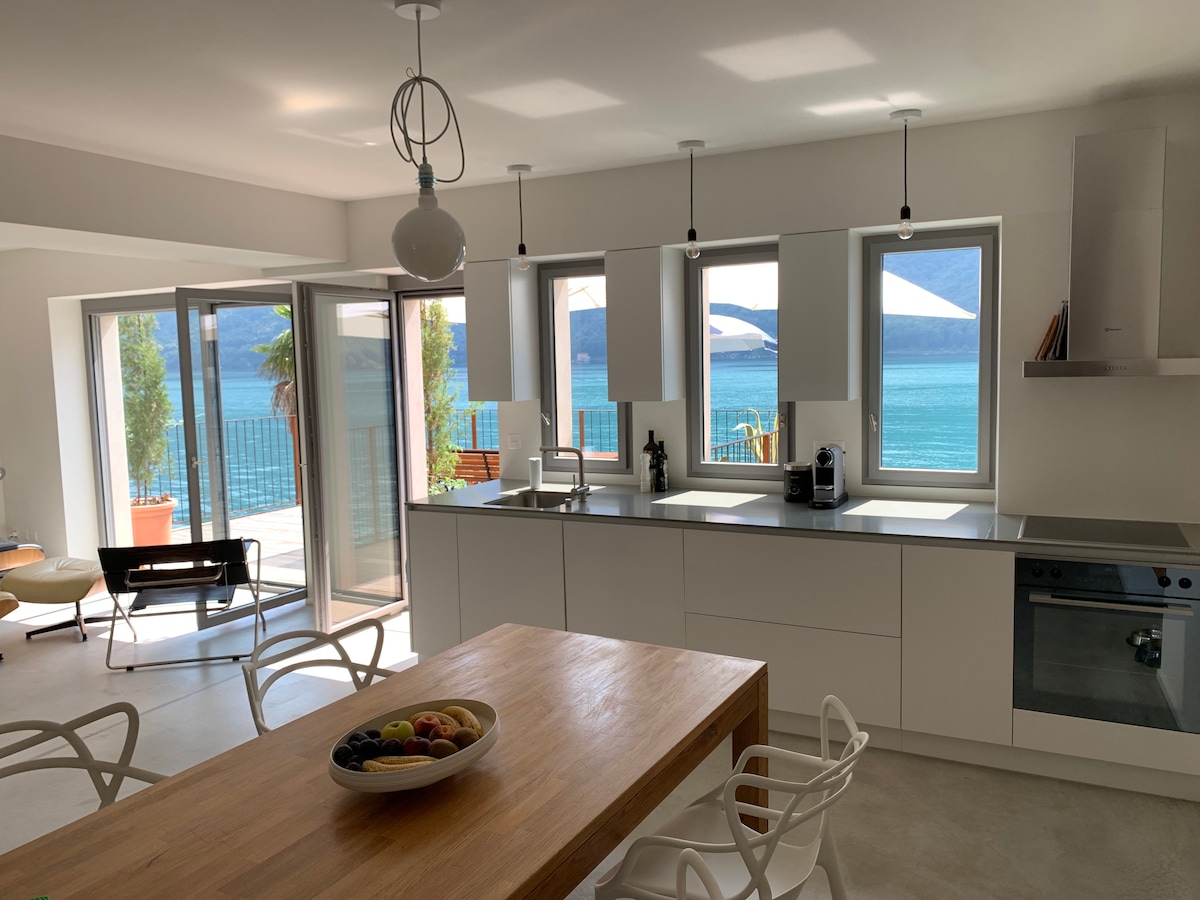 An open kitchen features a large wooden dining table surrounded by white chairs. Natural light floods the space through multiple windows, providing views of the lake. The modern kitchen includes sleek cabinetry and stainless-steel appliances, enhancing the contemporary design.