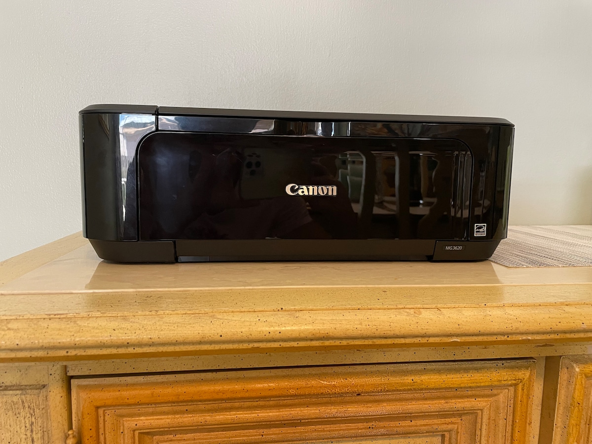 A black Canon printer is positioned on a wooden surface, showcasing a sleek and modern design. Its reflective surface allows light to create subtle highlights. The underlying furniture features a textured wood finish, complementing the printer's contemporary appearance.