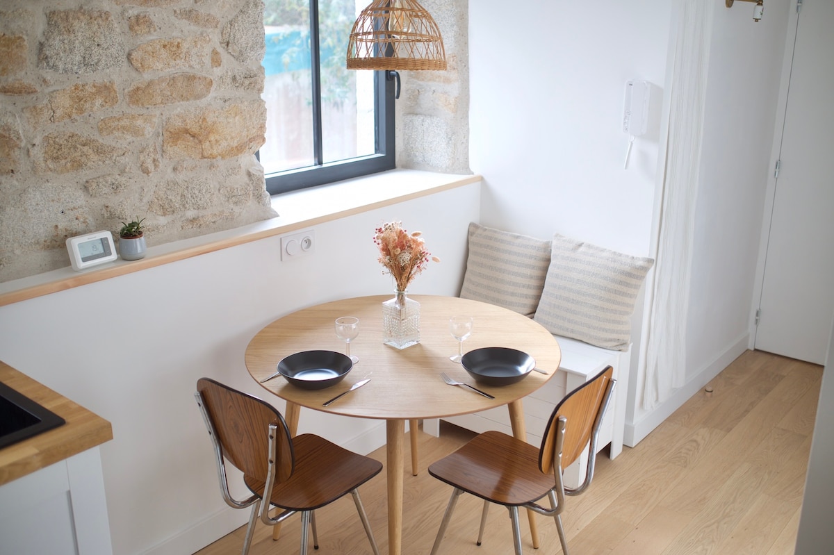 A dining area features a round wooden table surrounded by two chairs. A small vase with flowers rests at the center of the table, complemented by two plates and glasses. Natural light fills the space through a large window, highlighting the stone wall.