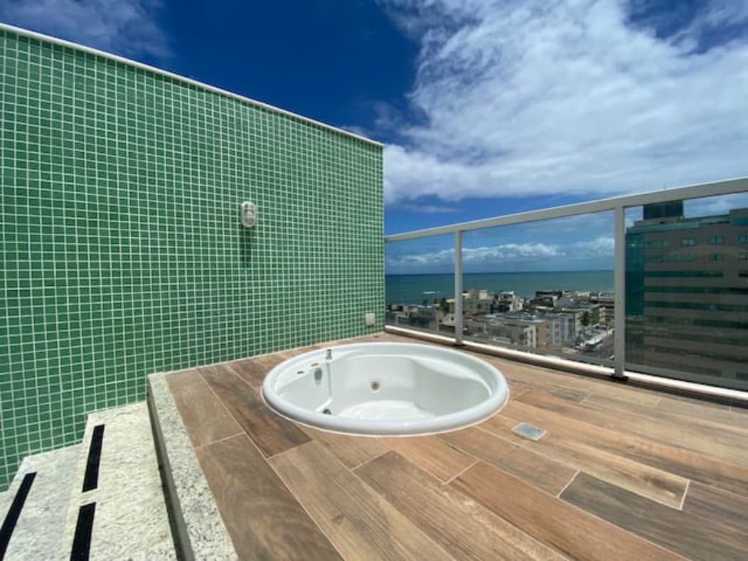 A private sun terrace is equipped with a circular jacuzzi, set against a backdrop of blue sky and coastal views. The surrounding area features green tiles and wooden decking, offering a serene space for relaxation.