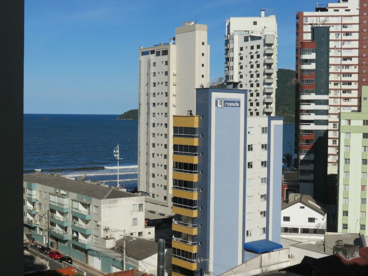 The view from the balcony shows a coastal scene featuring several high-rise buildings lining the beach. The ocean is visible in the background, with waves gently breaking against the shore. A clear blue sky complements the urban landscape.