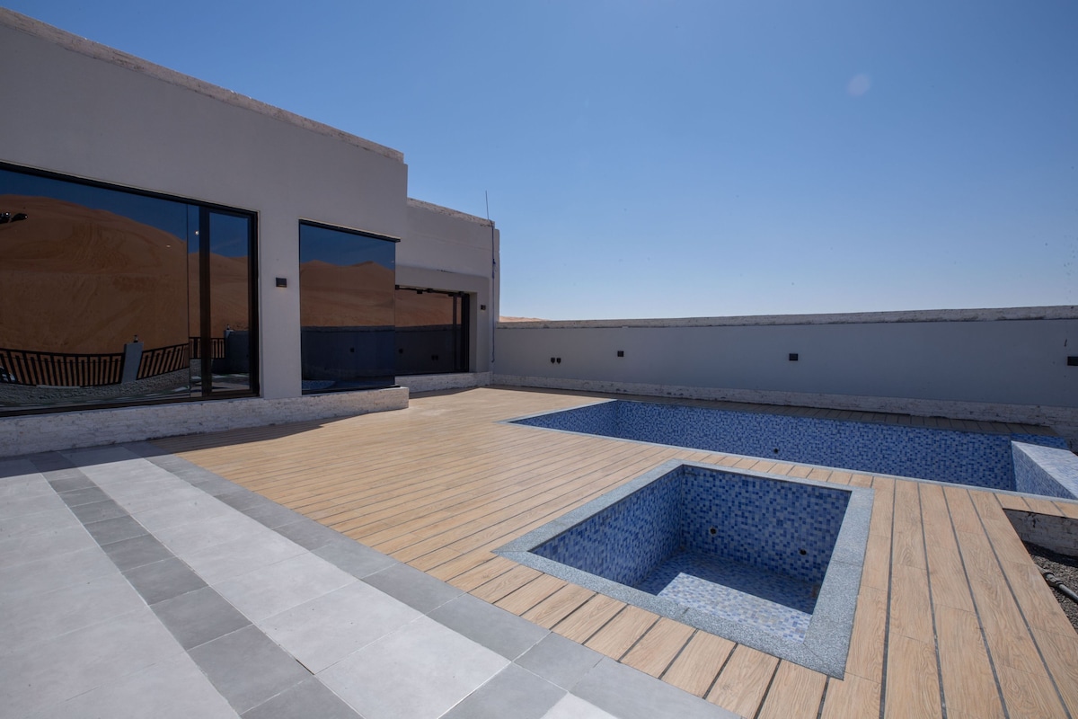 An expansive outdoor area features a wooden deck surrounding a mosaic-tiled pool and a smaller spa section. Large glass sliding doors provide access to the interior, with clear blue skies visible above.