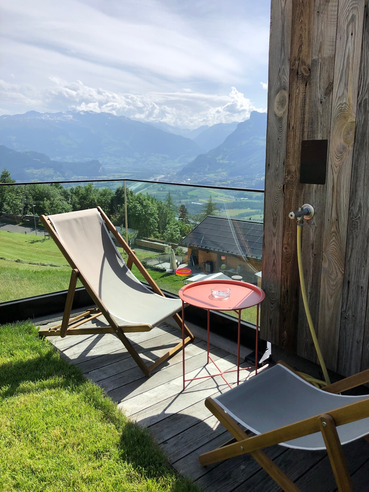 A pair of lounge chairs is positioned on a wooden deck, complemented by a small round table. A scenic mountain view, with lush greenery and distant peaks, is visible in the background under a clear sky.