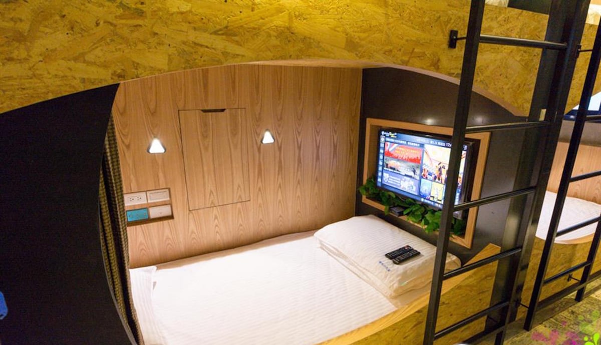 A cozy sleeping nook is presented, featuring a spacious bed with neatly arranged linens. Soft lighting is provided by mounted lamps on the wooden wall. A television and a small decorative plant are positioned on a shelf, enhancing the functional design of the space.