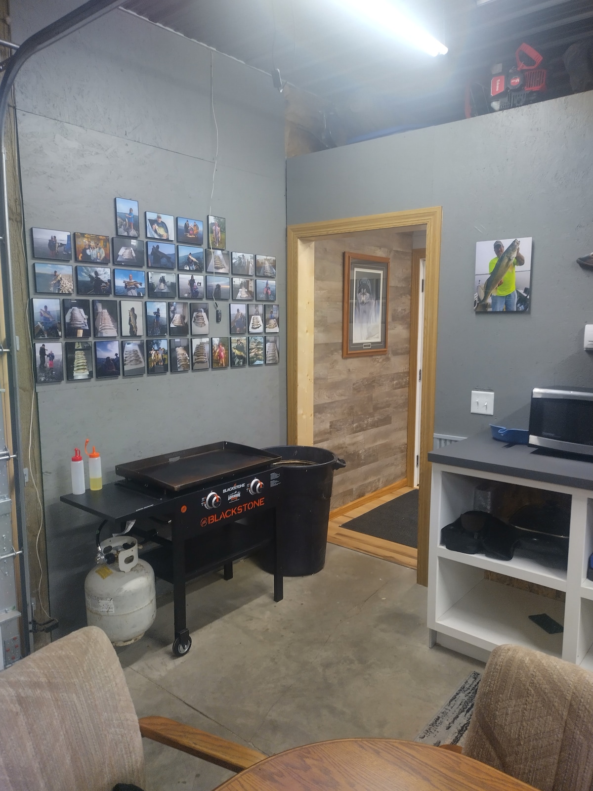 A functional cooking space features a Blackstone grill, positioned next to a wall adorned with various fishing-themed photographs. The doorway leads to another area, while a small countertop hosts an electric skillet and kitchen supplies. Soft lighting creates an inviting environment.