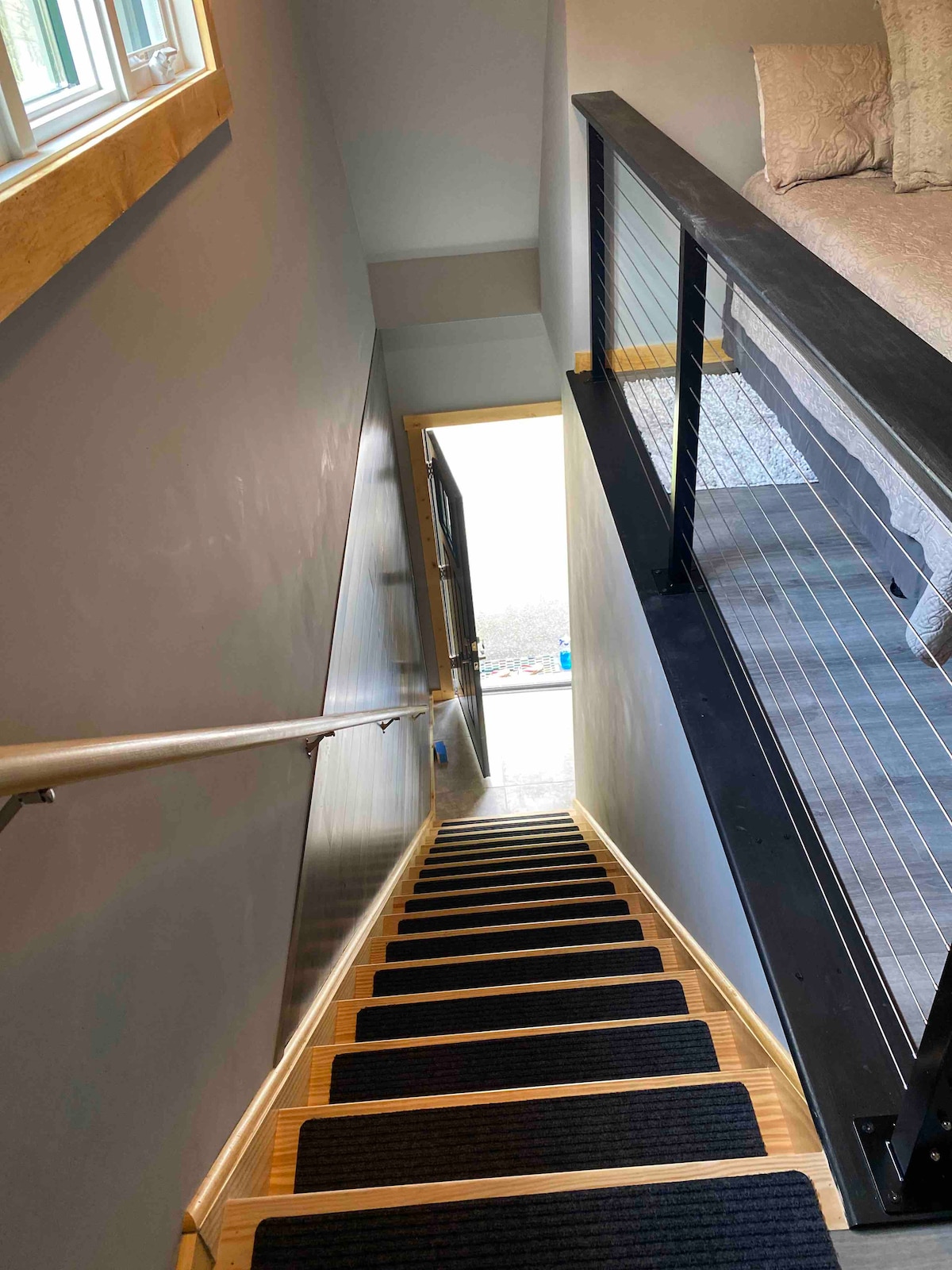 A staircase is visible, leading down from the loft to the entry door. The steps are covered with dark runners, and a handrail is positioned on one side. Natural light floods the space from a window at the bottom, illuminating the walls and floor.