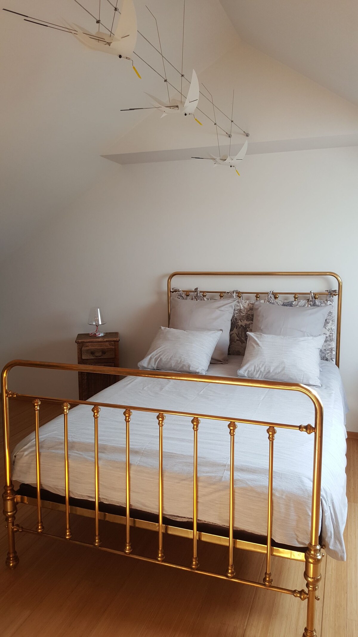 A bedroom features a light-colored wooden floor and a gold-framed bed with soft pillows. An accent table stands beside the bed. Delicate mobile decorations hang from the ceiling, adding a touch of whimsy to the minimalist space.