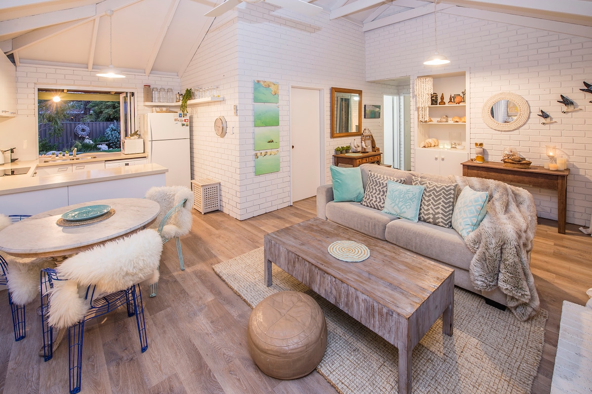 The open-plan living area features whitewashed brick walls and vaulted ceilings, with a cozy lounge area that includes a comfortable sofa and rustic coffee table. A round dining table with upholstered chairs is visible, alongside a modern kitchen equipped with essential appliances and ample natural light.