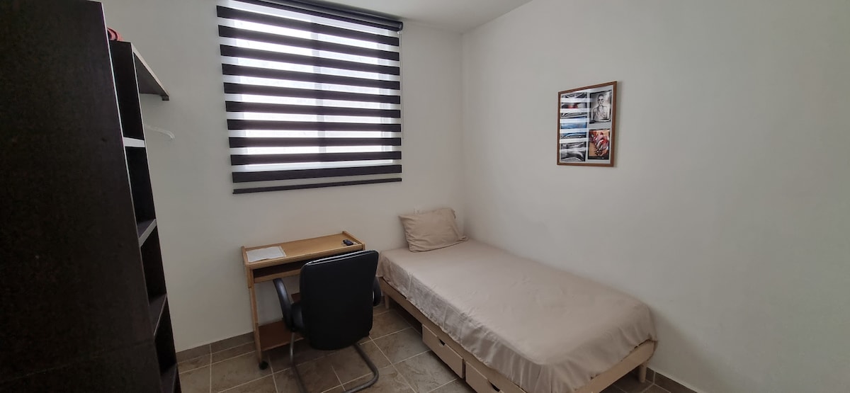 A simple room features a compact bed with a light-colored coverlet, positioned against a white wall. A wooden desk with a chair is set near the wall, with a framed image displayed above. A window is covered by horizontal striped blinds, allowing filtered light into the space.