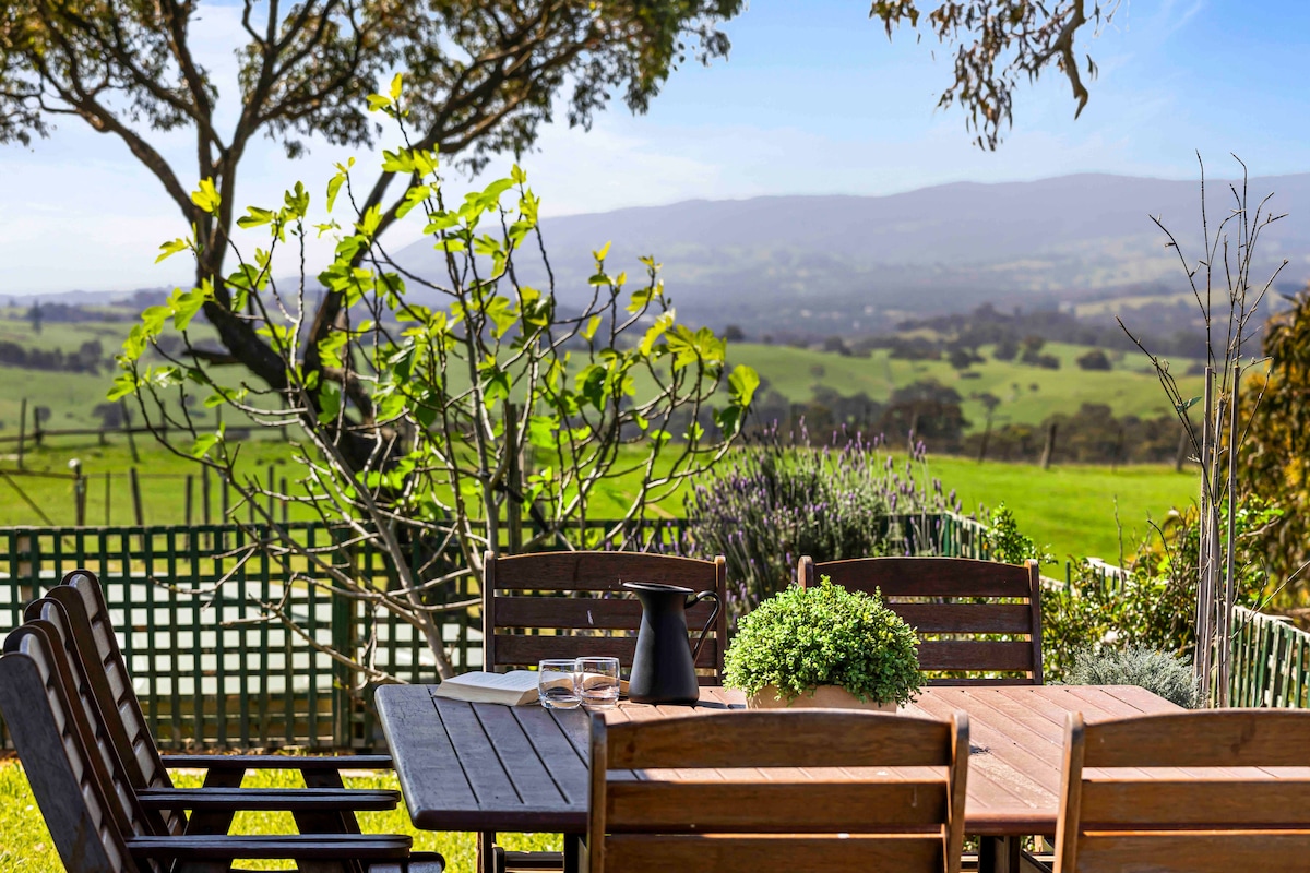 An outdoor dining area features a wooden table surrounded by several sturdy chairs. Lush greenery and blooming plants are visible in the foreground, while rolling hills stretch into the distance under a clear sky.