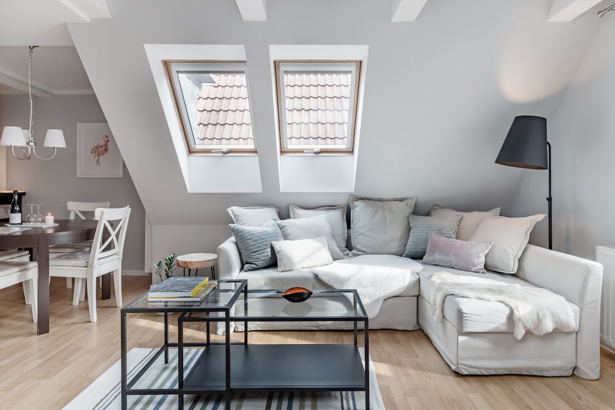 A bright living area features three skylight windows, allowing natural light to fill the space. A spacious light-colored sofa is complemented by various textured cushions, and a low coffee table rests on a striped area rug. A dining table with white chairs is positioned nearby.