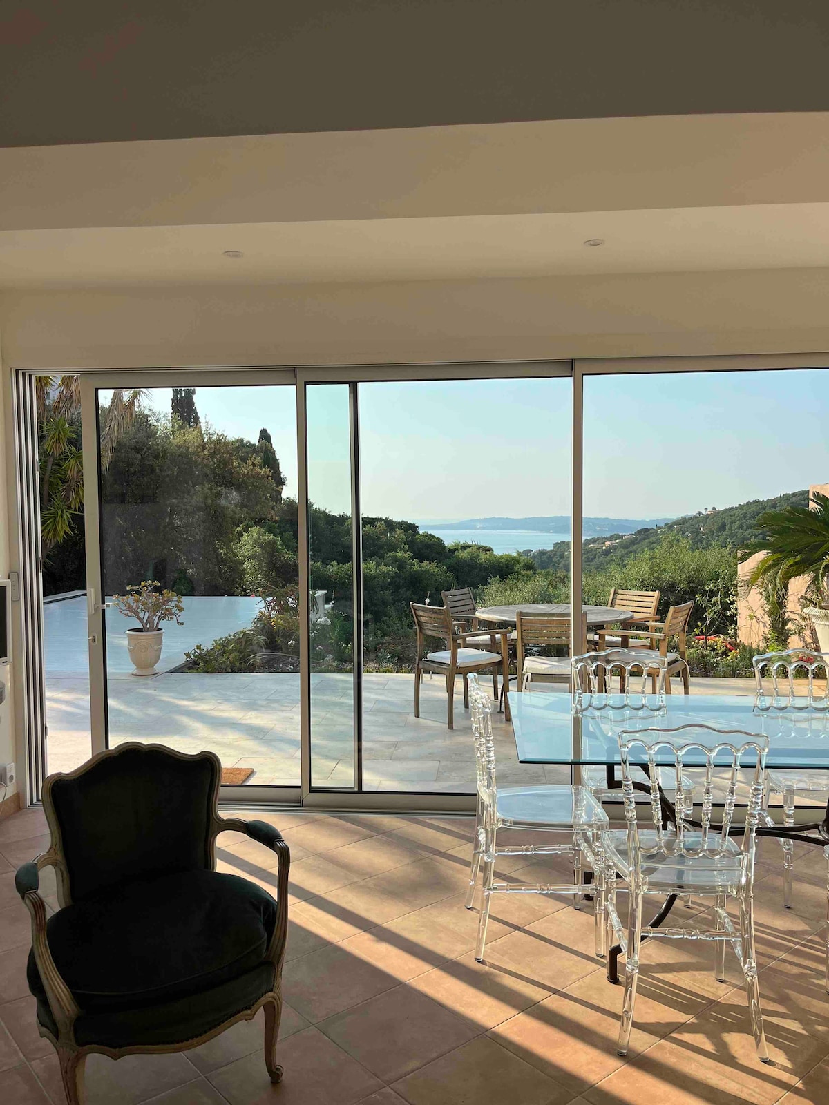 Large glass doors open to reveal a scenic view of the surrounding landscape and water. A clear dining table and chairs are positioned indoors, while outdoor seating is visible on the terrace, which is enhanced by lush greenery.